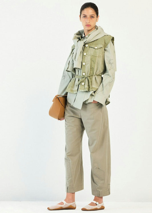 A model stands against a plain white studio background wearing a light olive green ruffled utility jacket over a pale sage green top, paired with matching light olive trousers and white ballet flats, and holding a small tan shoulder bag.