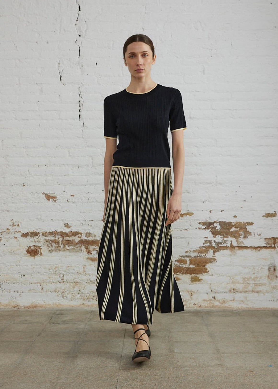 Woman wearing a black top and striped skirt against a white brick wall.