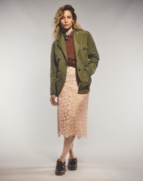 A model stands against a light gray studio background wearing an olive green utility jacket over a rust-brown collared shirt, paired with a light pink textured midi skirt and dark brown leather shoes.