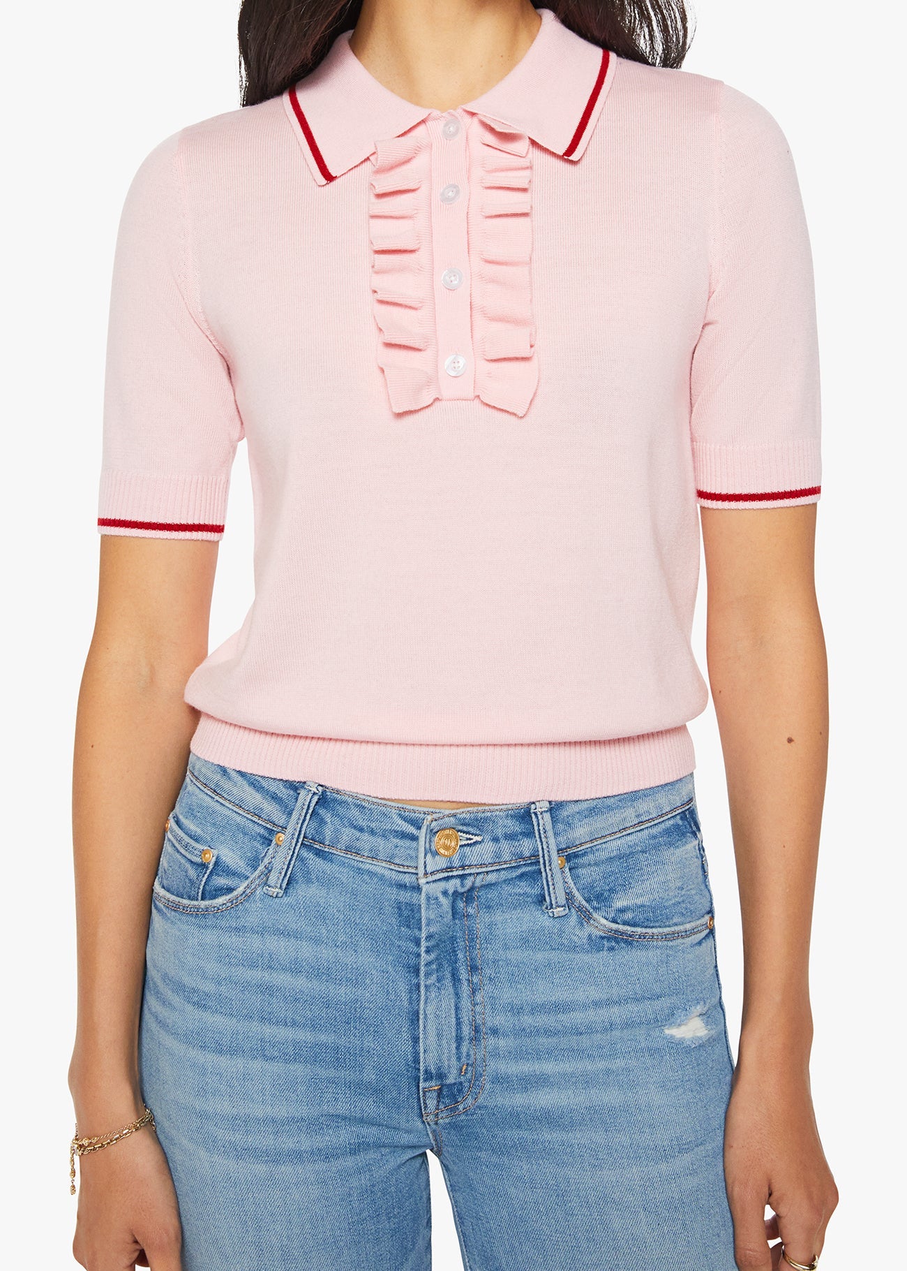 A woman wearing a light pink short-sleeve knit polo shirt with red piping trim on the collar, sleeve cuffs, and a ruffled placket detail, paired with blue jeans.
