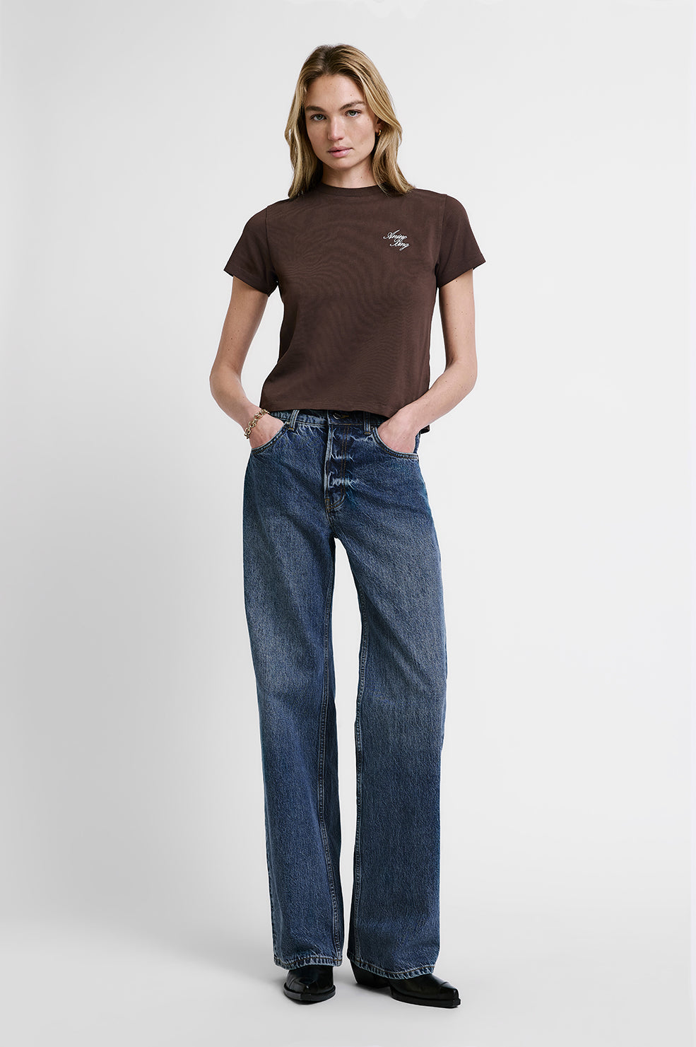 A woman modeling a brown short-sleeve crewneck t-shirt with a small white chest graphic, paired with high-waisted, wide-leg blue jeans and black shoes, against a light gray studio background.