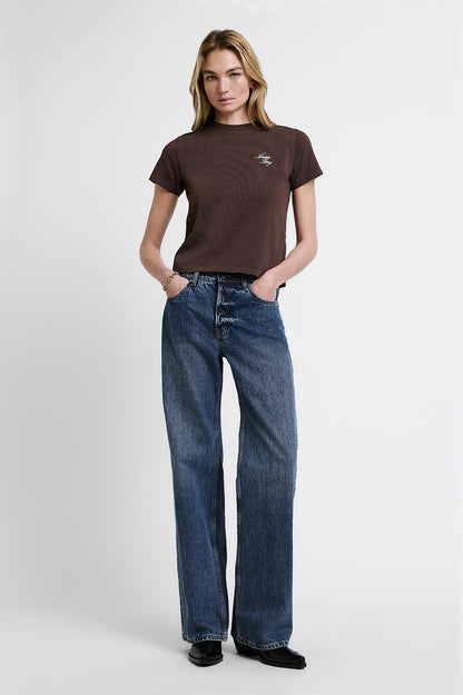 A woman modeling a brown short-sleeve crewneck t-shirt with a small white chest graphic, paired with high-waisted, wide-leg blue jeans and black shoes, against a light gray studio background.