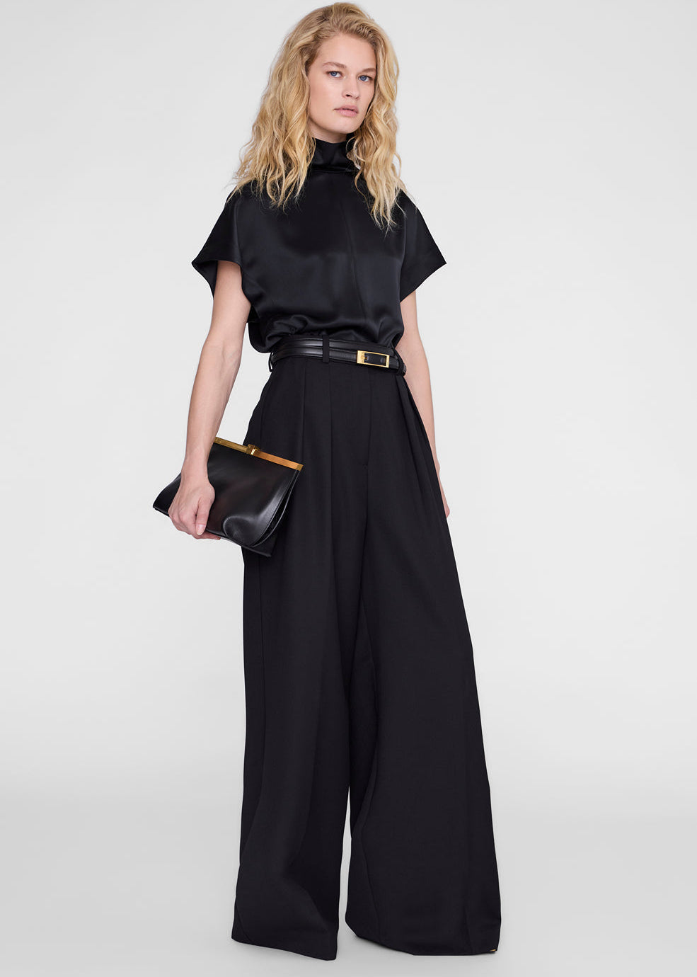 A model wearing a black satin short-sleeve mock-neck top and matching wide-leg trousers, styled with a black belt and a black clutch bag with gold trim, against a light gray studio background.