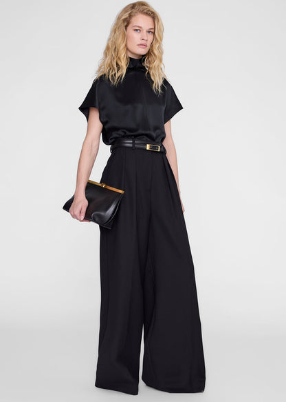 A model wearing a black satin short-sleeve mock-neck top and matching wide-leg trousers, styled with a black belt and a black clutch bag with gold trim, against a light gray studio background.