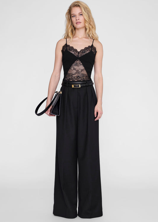 A blonde model stands against a light gray studio background wearing a black lace-trimmed camisole top with matching wide-leg trousers, a black belt with a gold buckle, and holding a black handbag.