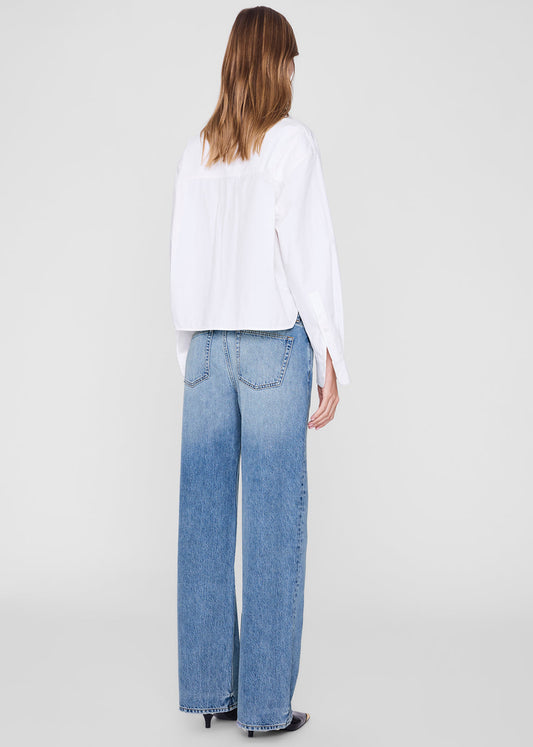 A woman shown from the back wearing a white long-sleeve blouse and wide-leg light blue jeans against a plain light gray studio background.
