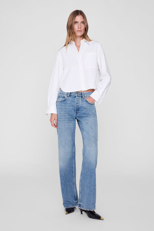 A woman modeling a white long-sleeve button-up shirt and light-wash blue jeans against a plain light gray studio background.