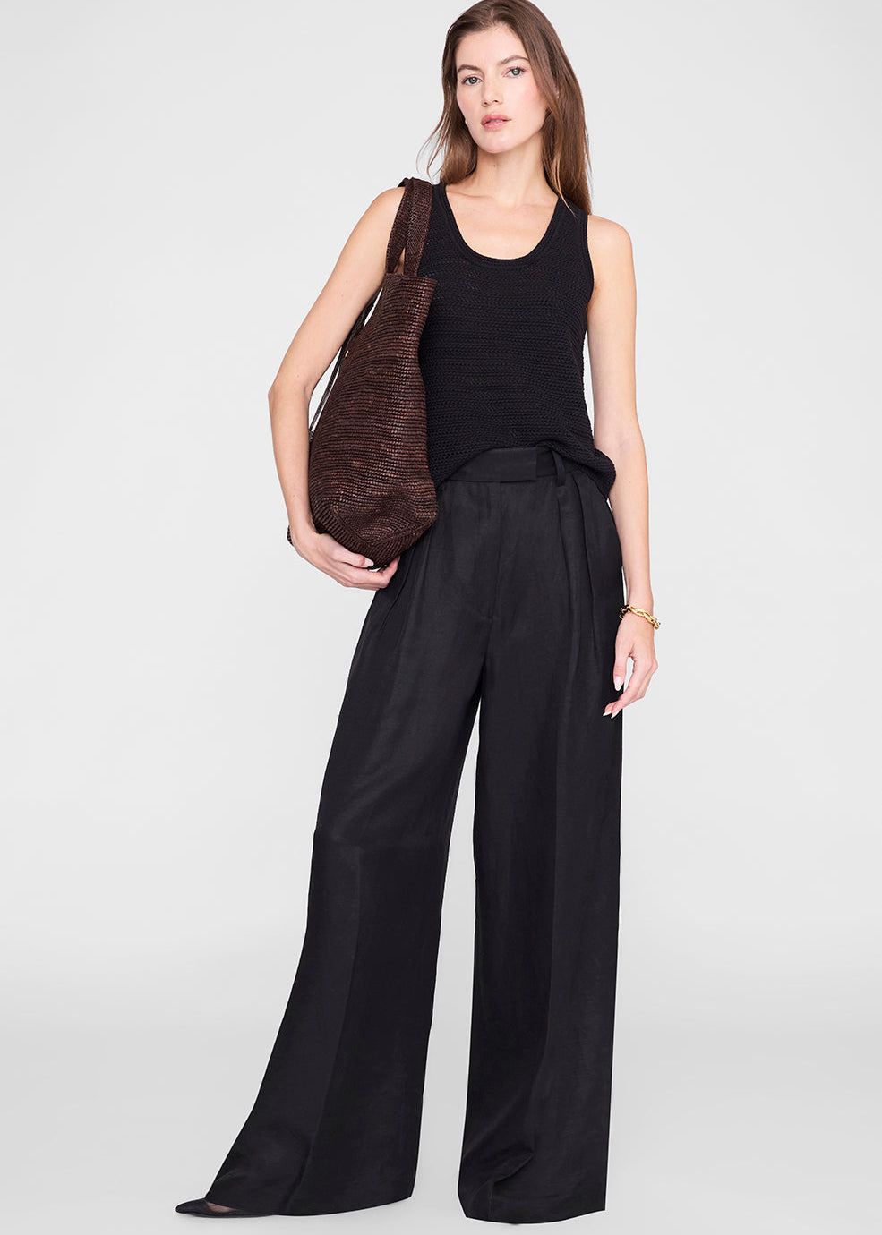 Woman wearing a black sleeveless top and wide-leg pants, holding a brown bag on a white background