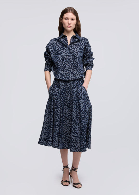 A woman modeling a dark navy blue and black leopard-print shirt and skirt set with black strappy heels, shot against a solid light gray background.