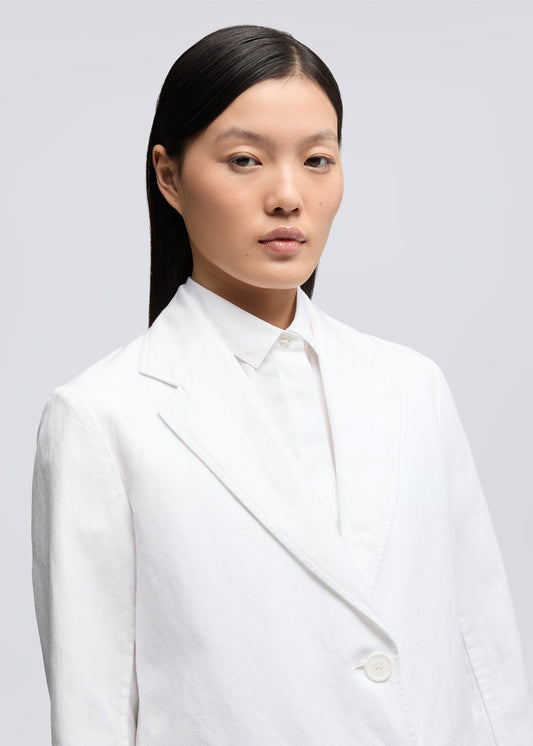 A model with long, straight black hair wearing a white double-breasted blazer over a white collared shirt, photographed against a solid light gray background.
