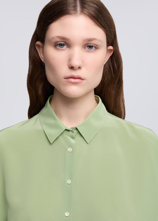 A close-up shot of a model wearing a light sage green button-down shirt with a pointed collar, set against a solid light gray background.