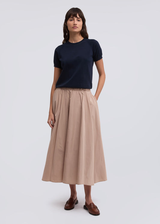 A woman stands against a light gray studio background wearing a dark navy short-sleeve crewneck top, a pleated beige midi skirt, and brown loafers.