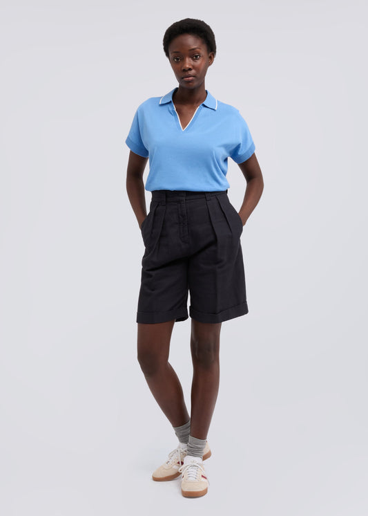 A model stands against a light gray studio background wearing a light blue short-sleeve polo shirt tucked into dark charcoal pleated shorts, paired with gray socks and beige sneakers.