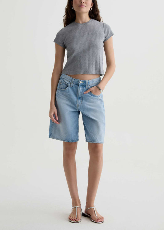 A model wearing a gray short-sleeve cropped knit top and light-wash blue denim shorts, paired with silver thong sandals, standing against a plain off-white studio background.
