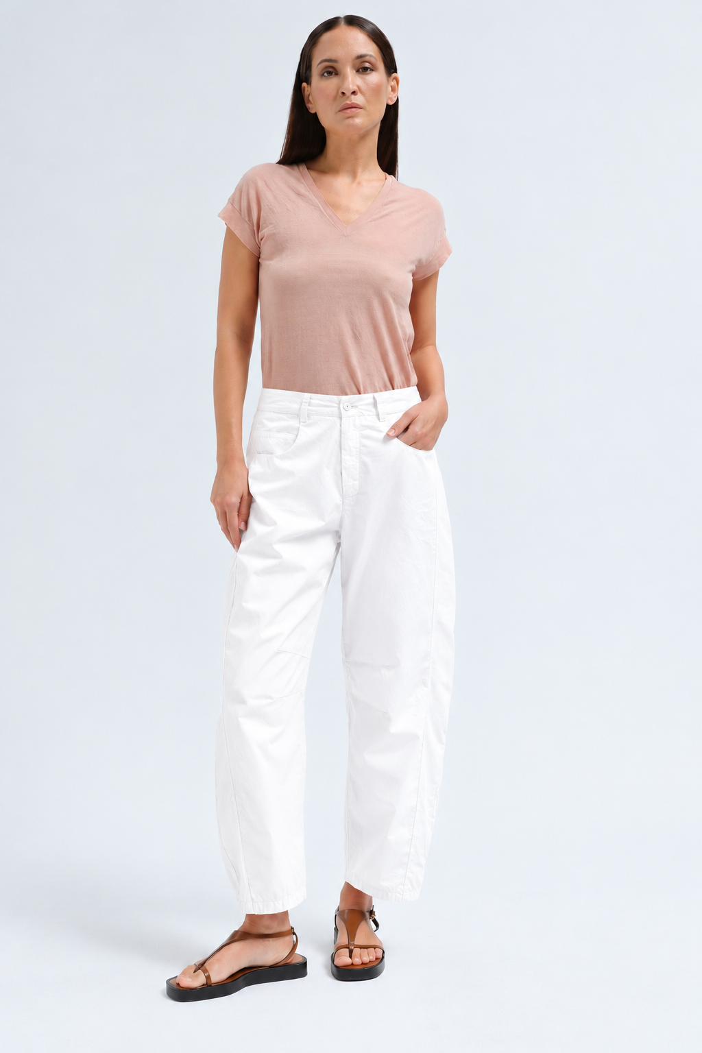A woman modeling a light pink short-sleeve V-neck t-shirt and white cropped pants with black sandals, standing against a light gray studio background.