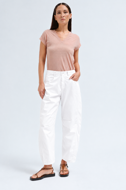A woman modeling a light pink short-sleeve V-neck t-shirt and white cropped pants with black sandals, standing against a light gray studio background.