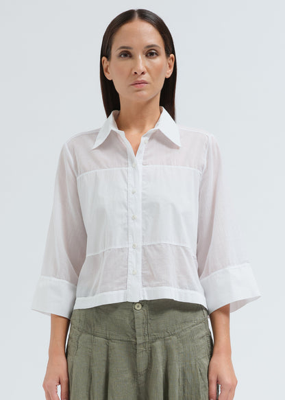 A woman modeling a white semi-sheer three-quarter sleeve button-up blouse with a collar and gathered front, paired with olive green pants, against a light gray studio background.