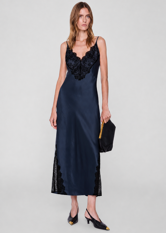 A woman models a navy blue satin slip dress with black lace trim on the bodice and hem, styled with black slingback heels and a black clutch bag against a light gray studio background.