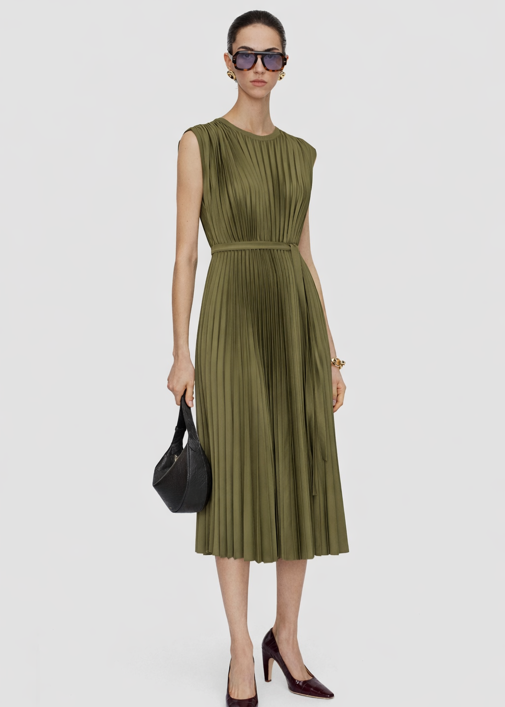 Woman wearing a green pleated dress with sunglasses and a black handbag on a white background