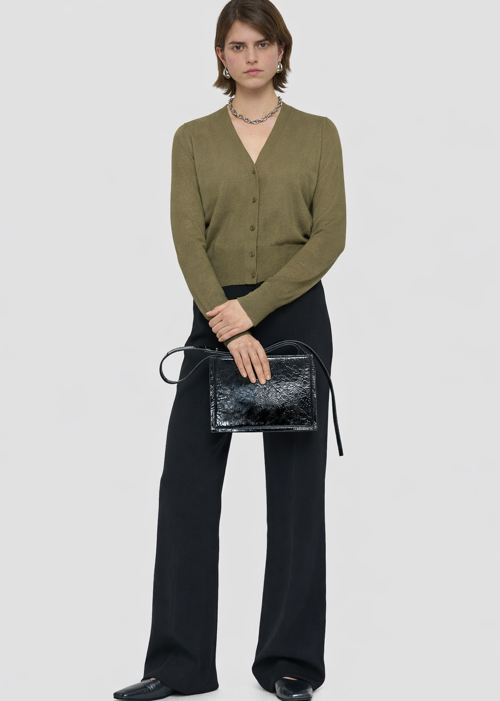 Woman holding a black handbag wearing a green cardigan and black pants on a white background