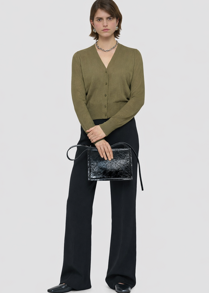 Woman holding a black handbag wearing a green cardigan and black pants on a white background
