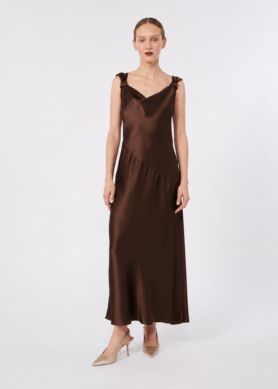 A woman modeling a sleeveless, V-neck brown satin maxi dress with shoulder straps, paired with beige high heels, against a light gray studio background.
