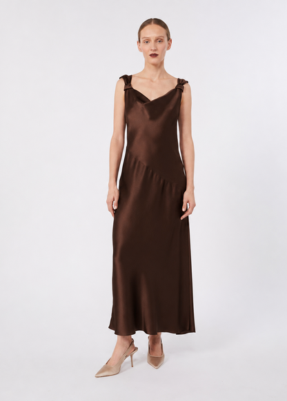 A woman modeling a sleeveless, V-neck brown satin maxi dress with shoulder straps, paired with beige high heels, against a light gray studio background.