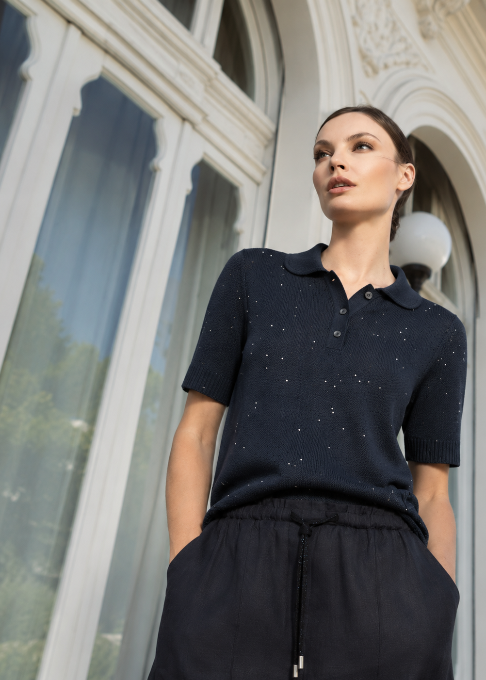 A woman wearing a dark navy short-sleeve knit polo shirt with tiny silver speckles and black drawstring trousers, standing in front of a large white arched window.