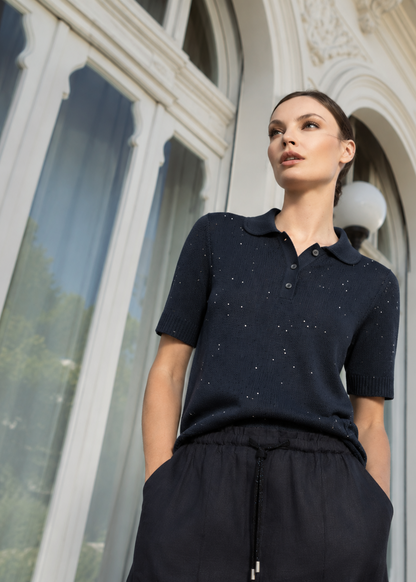 A woman wearing a dark navy short-sleeve knit polo shirt with tiny silver speckles and black drawstring trousers, standing in front of a large white arched window.