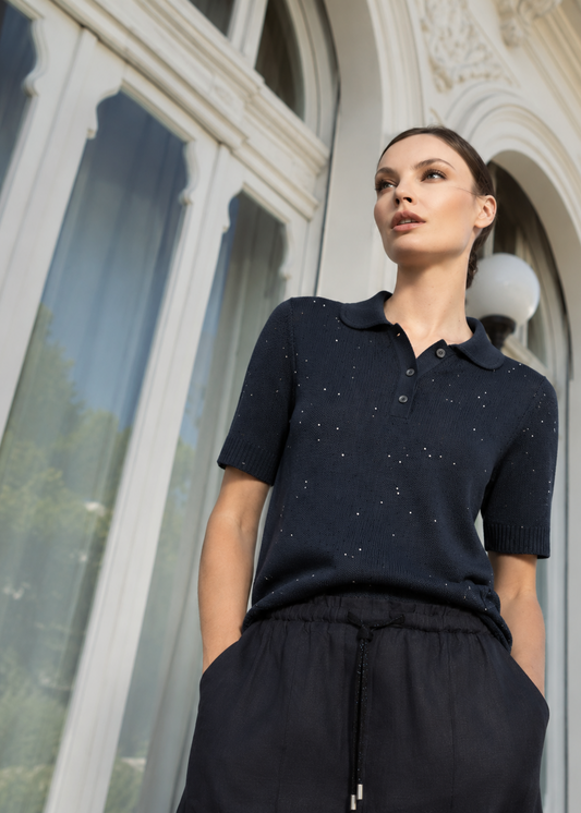 A woman wearing a dark navy short-sleeve knit polo shirt with tiny silver speckles and black drawstring trousers, standing in front of a large white arched window.