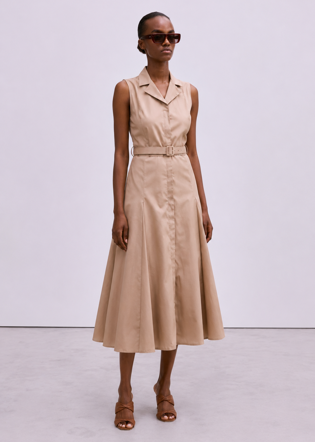 A model wearing a sleeveless beige midi dress with a collar, button front, and matching belt, styled with brown strappy sandals and dark sunglasses against a light gray studio background.
