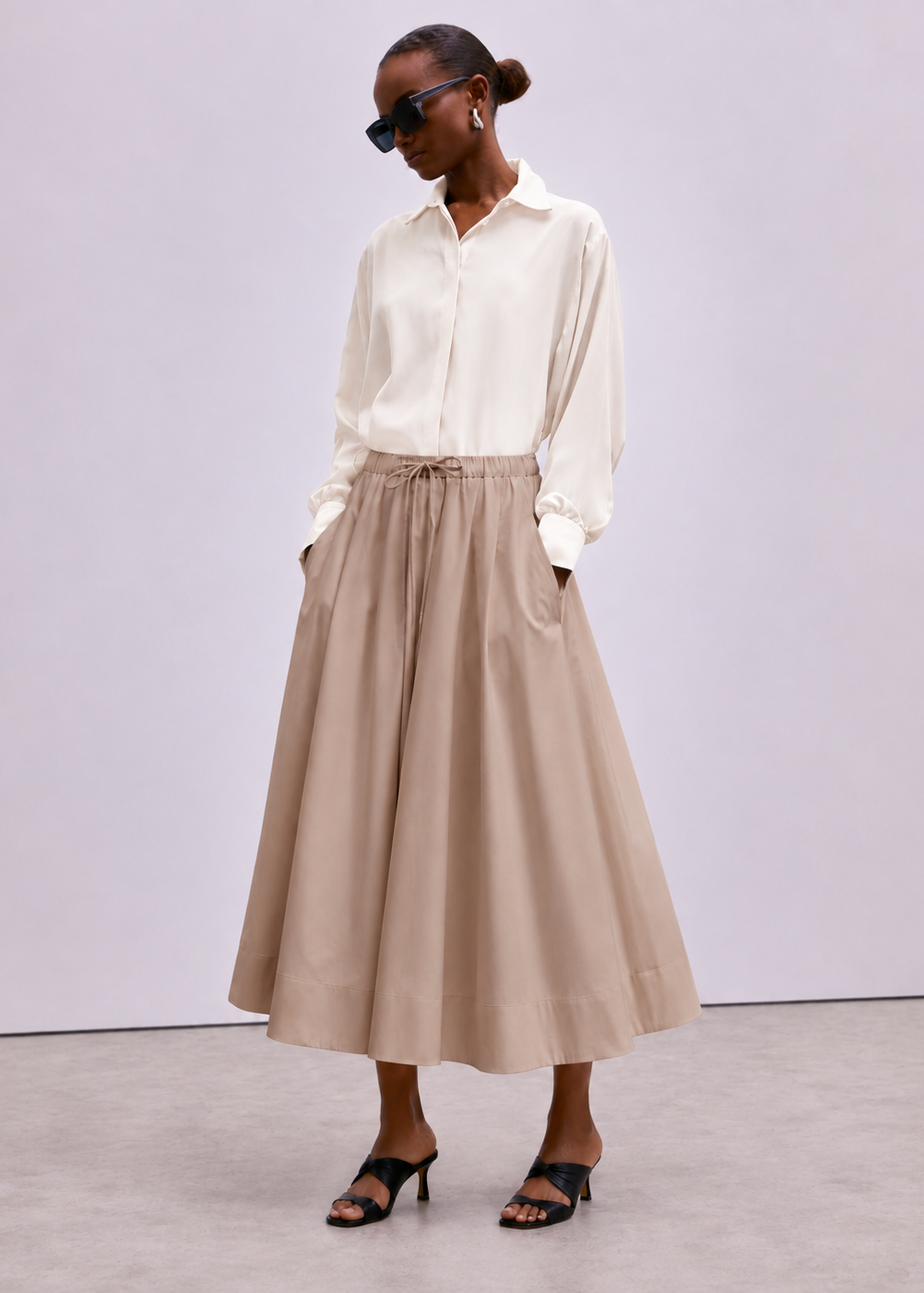 A model wearing a cream long-sleeve button-up shirt and a beige high-waisted midi skirt with a drawstring waist, styled with black sunglasses and black heeled sandals against a light gray studio background.