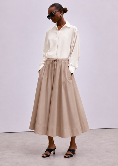 A model wearing a cream long-sleeve button-up shirt and a beige high-waisted midi skirt with a drawstring waist, styled with black sunglasses and black heeled sandals against a light gray studio background.