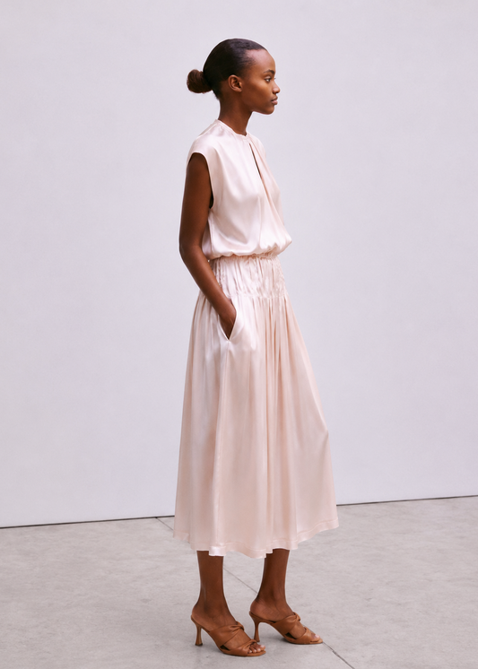 A woman stands in profile wearing a sleeveless light blush pink midi dress with a gathered waist and side pockets, paired with tan heeled sandals, against a plain light gray studio background.