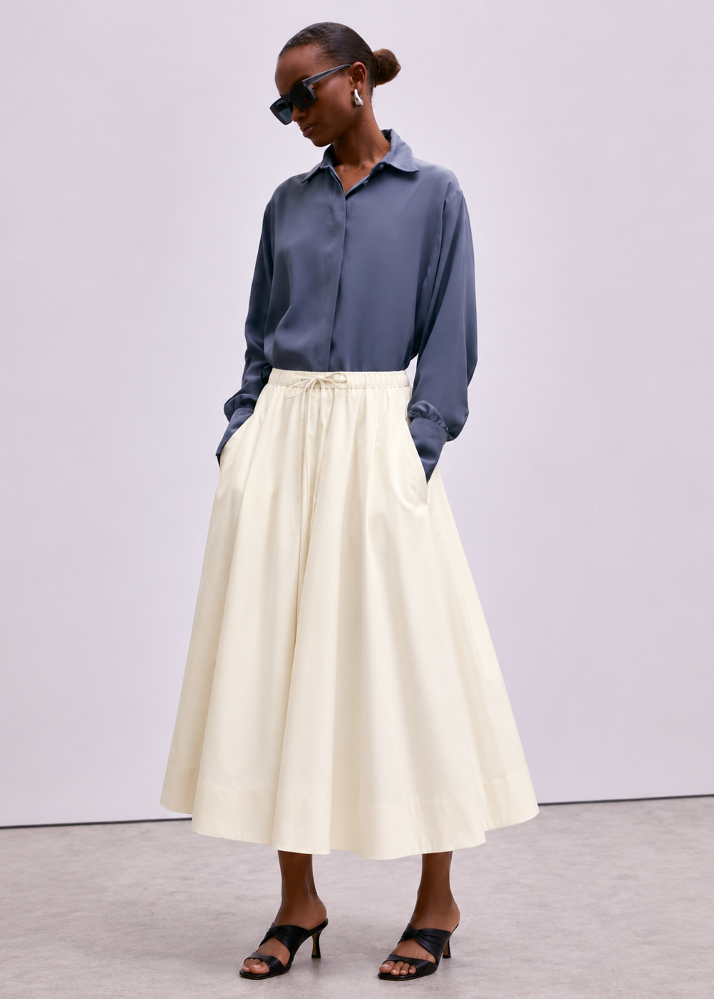 A model wearing a slate blue long-sleeve button-up shirt and a high-waisted cream A-line midi skirt with a drawstring waist, paired with black strappy heels, against a neutral studio backdrop.