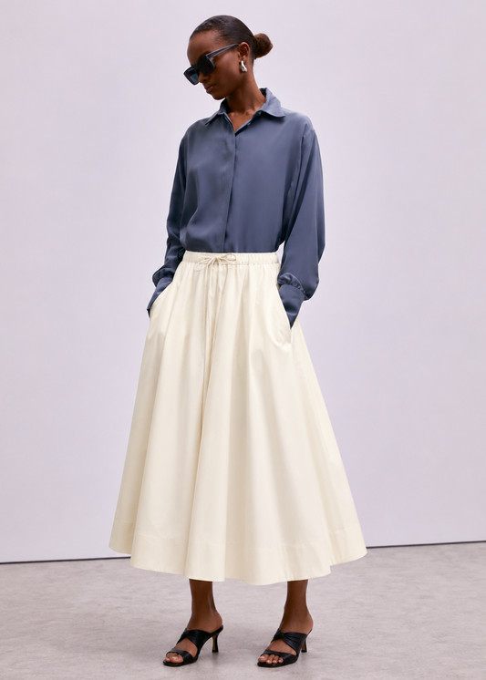 A model wearing a slate blue long-sleeve button-up shirt and a high-waisted cream A-line midi skirt with a drawstring waist, paired with black strappy heels, against a neutral studio backdrop.