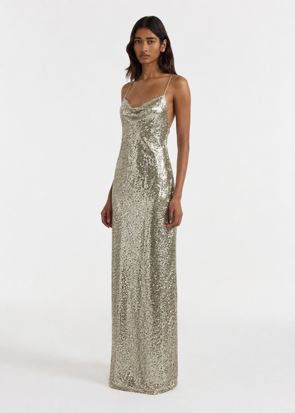 A model wearing a floor-length, spaghetti-strap sequined gown in a shimmering champagne gold color, photographed against a plain white studio background.