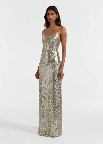 A model wearing a floor-length, spaghetti-strap sequined gown in a shimmering champagne gold color, photographed against a plain white studio background.