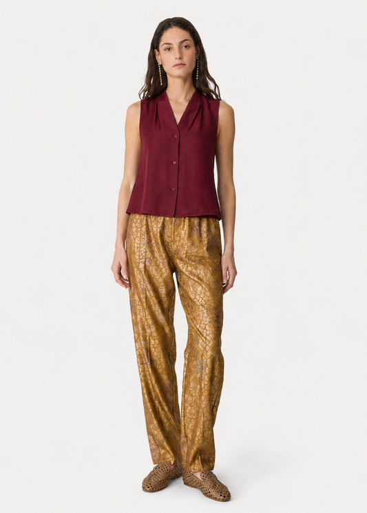 A model stands against a light gray studio background wearing a sleeveless burgundy top and shiny gold patterned pants.