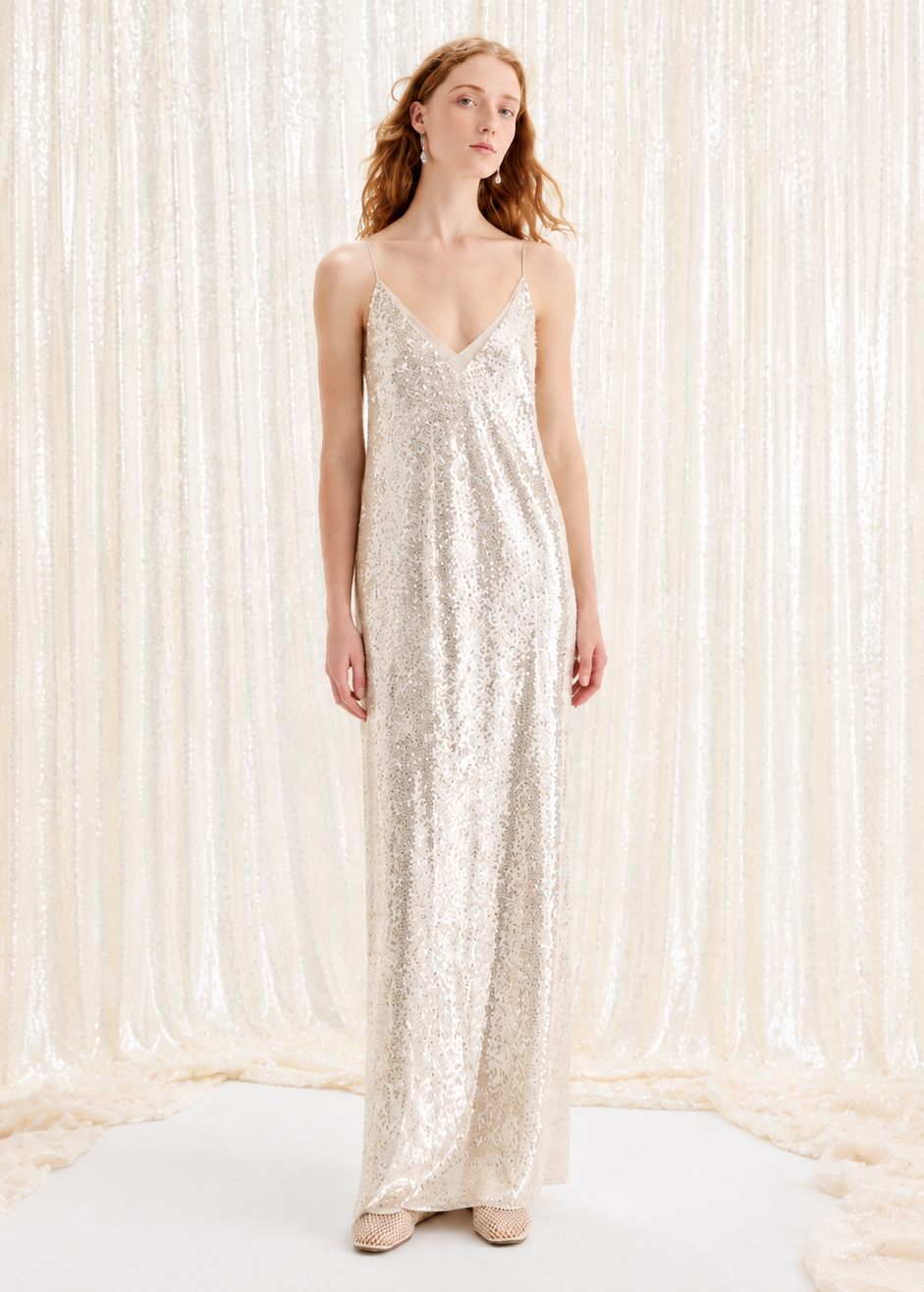 A woman stands in a long, floor-length champagne sequin slip dress with thin straps, posed against a shimmering cream curtain backdrop.