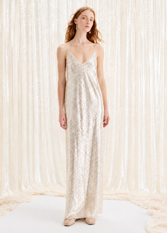 A woman stands in a long, floor-length champagne sequin slip dress with thin straps, posed against a shimmering cream curtain backdrop.