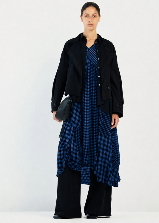 A model stands against a plain white studio background wearing a black cropped jacket over a blue and black checkered dress, paired with wide-leg black pants and holding a small black handbag.