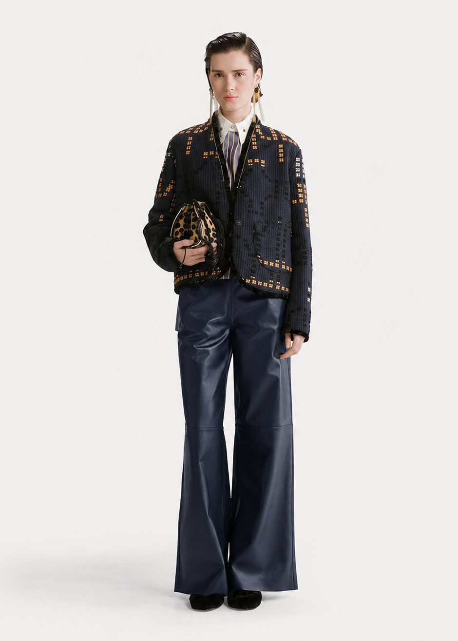 A model stands against a light gray studio background wearing a dark embroidered jacket with gold-toned embellishments, dark wide-leg pants, a white shirt, and a patterned tie, while holding a small patterned handbag.
