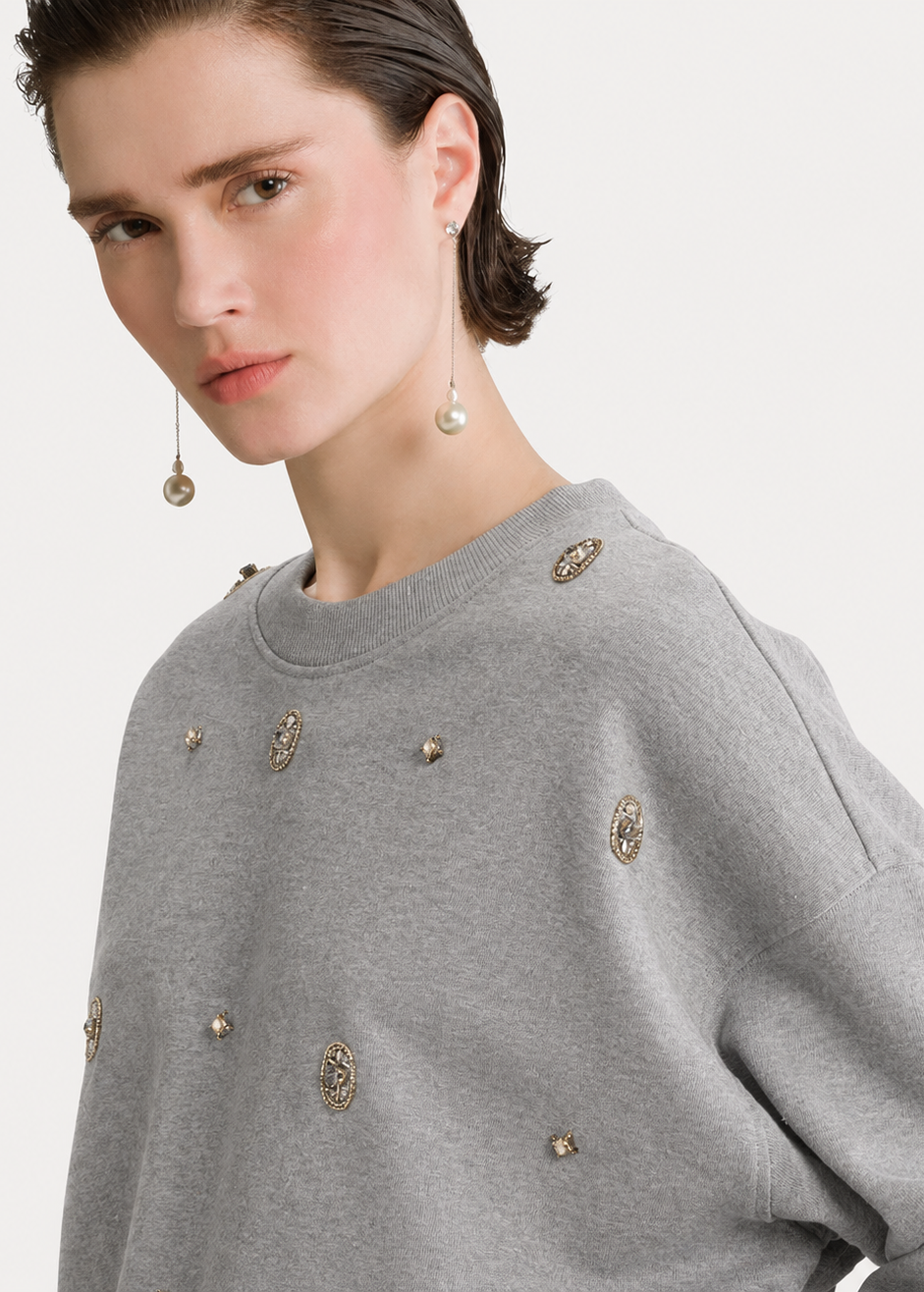 A close-up of a model wearing a light gray crewneck sweatshirt decorated with metallic gold and silver ornate studs, set against a plain light gray background.