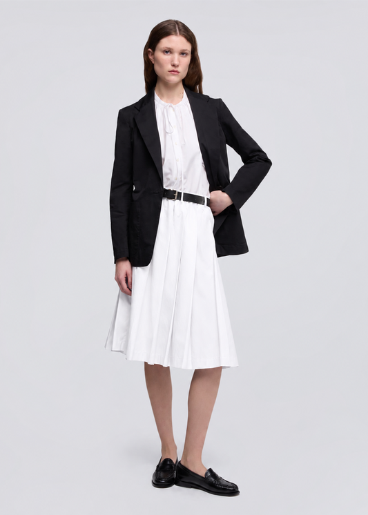 A woman modeling a black blazer over a white pleated midi dress with a black belt, paired with black loafers, against a light gray studio background.