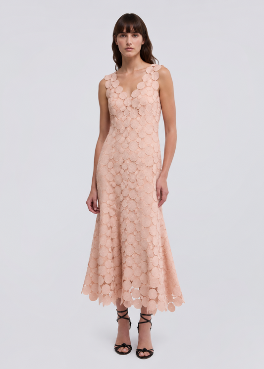 A woman modeling a sleeveless, floor-length peach lace dress with 3D floral applique, paired with black strappy high-heeled sandals, against a light gray studio background.