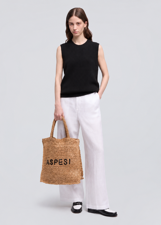A model stands against a light gray studio background wearing a black sleeveless top, white wide-leg trousers, and black loafers, while holding a natural woven straw tote bag with the brand name 'ASPESI' printed in black.