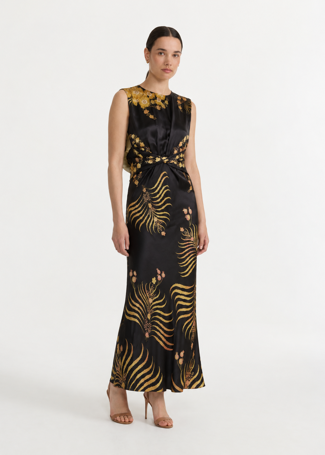 A woman modeling a sleeveless black satin maxi dress with a gold botanical and fern-like pattern, styled with nude high-heeled sandals against a plain white studio background.