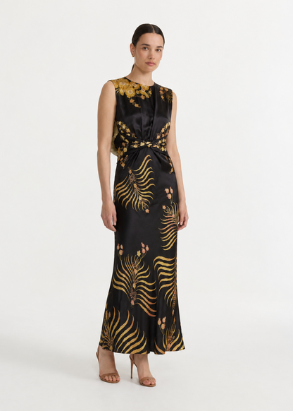 A woman modeling a sleeveless black satin maxi dress with a gold botanical and fern-like pattern, styled with nude high-heeled sandals against a plain white studio background.