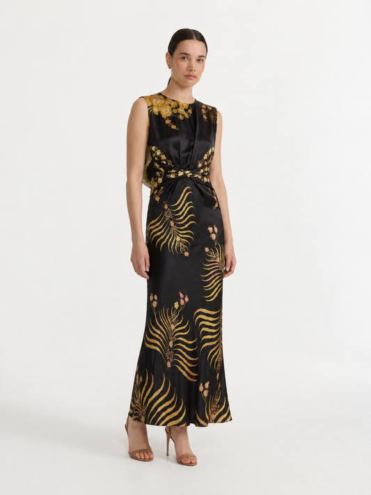 A woman modeling a sleeveless black satin maxi dress with a gold botanical and fern-like pattern, styled with nude high-heeled sandals against a plain white studio background.